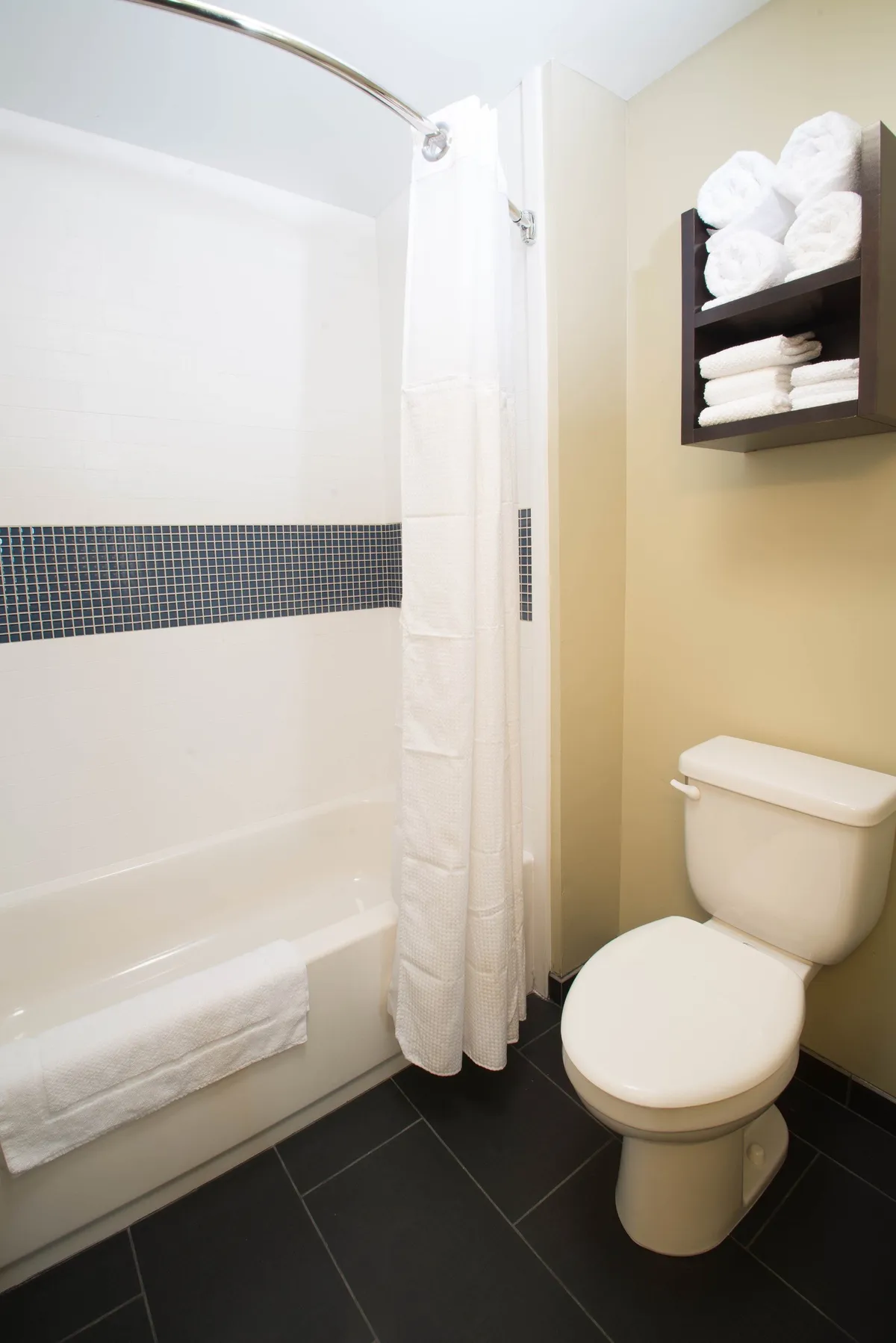 A modern bathroom equipped with a full bathtub, shower curtain, toilet, and a neatly organized rack of plush towels. Clean shelves display complimentary toiletries, reinforcing a sense of care and convenience. The space exudes freshness and modernity.