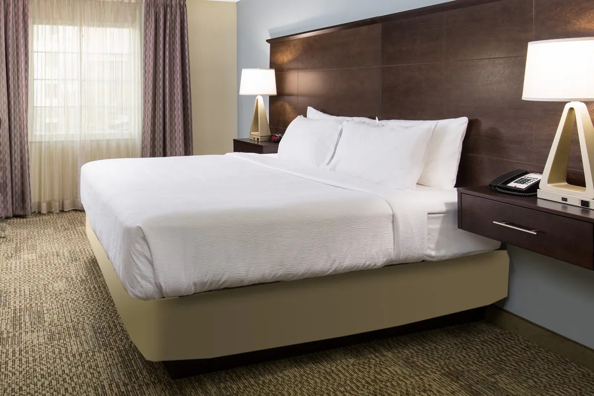 A spacious bedroom featuring a plush king bed topped with crisp white linens. Soft natural light pours in through a large window, highlighting the calming neutral tones of the space. A nightstand with lamps and a cozy reading nook evoke an atmosphere of restful privacy and comfort.