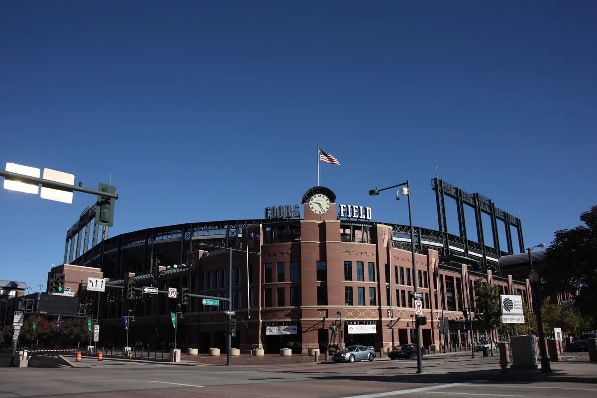 The exterior view of Ford Field, featuring the distinct stadium structure with an American flag in the foreground and vibrant activity around the main entrance. The scene includes families and groups heading toward the venue, surrounded by busy traffic signals and signs of a lively urban environment.