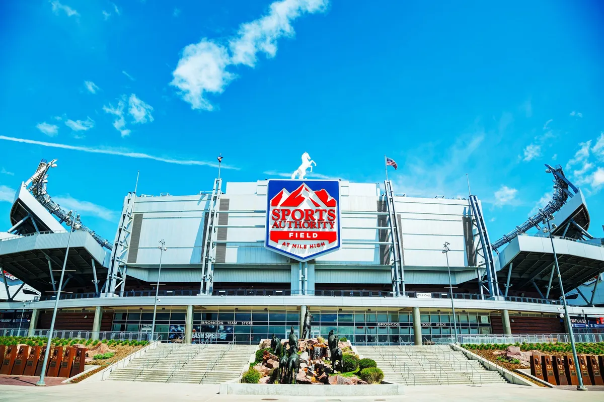 The image showcases the vibrant exterior of a bustling stadium, with its prominent facade and large logo clearly visible. Surrounded by well-maintained landscaping, the scene pulses with the energy of fans gathering for a major sports event or concert, encapsulating Denver’s passion for live entertainment and community gatherings.