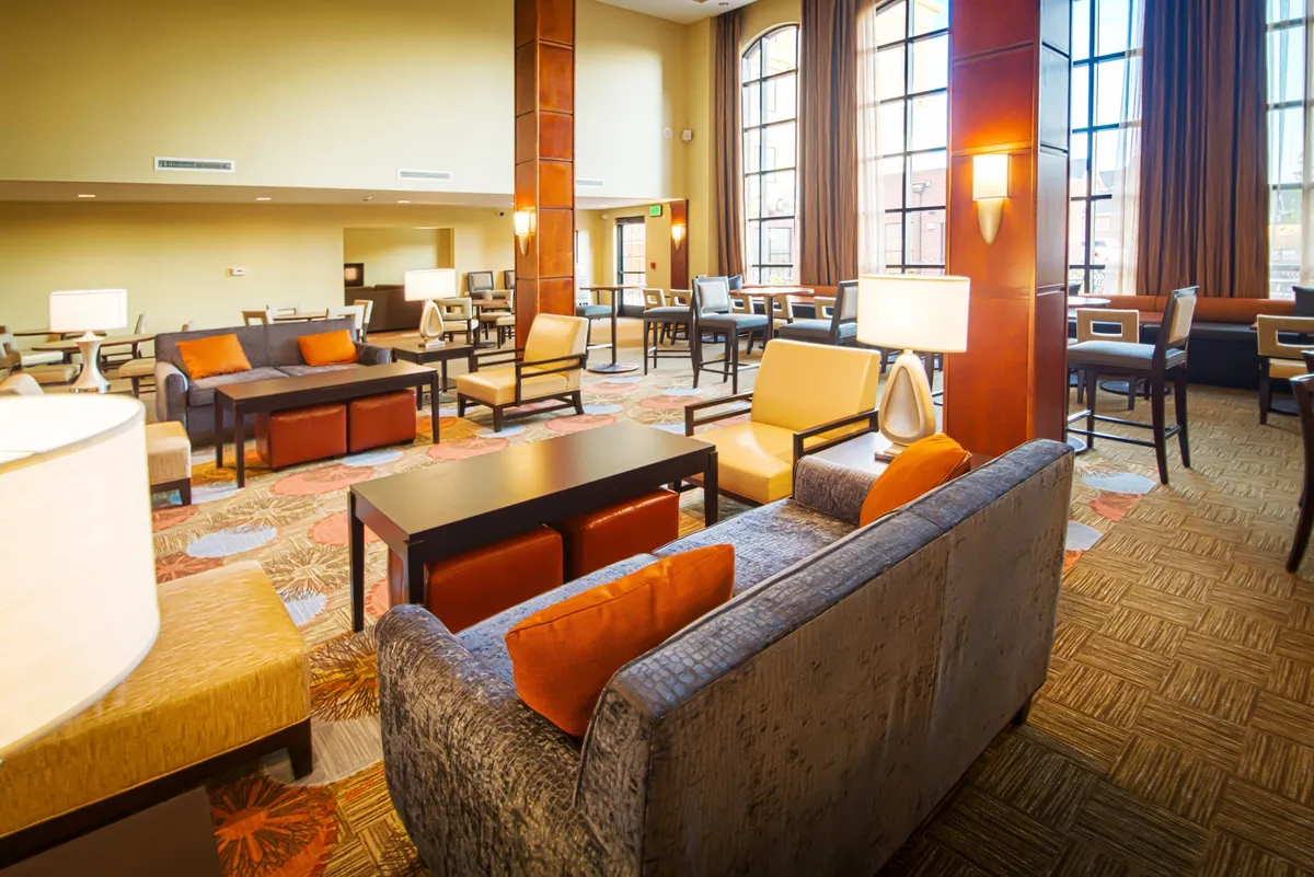 A sunlit living space featuring large windows that let in ample natural light, comfortable seating options arranged for both relaxation and small gatherings, a colorful modern rug, and well-placed lamps that create a cozy yet functional ambiance. The layout appears ideal for unwinding, hosting guests, or quiet moments alone.
