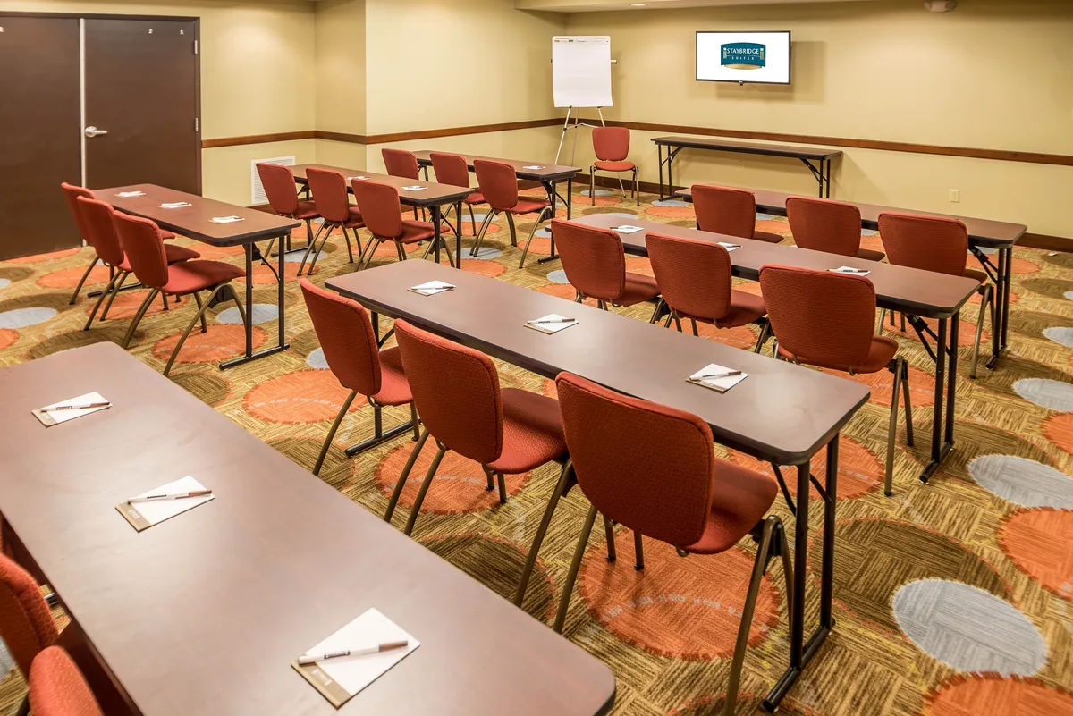 A professional business center set up with rows of tables and chairs, vibrant carpeting, a large presentation screen, and a flip chart. The space is well-lit and designed for collaboration, suitable for meetings, workshops, and other productive activities.