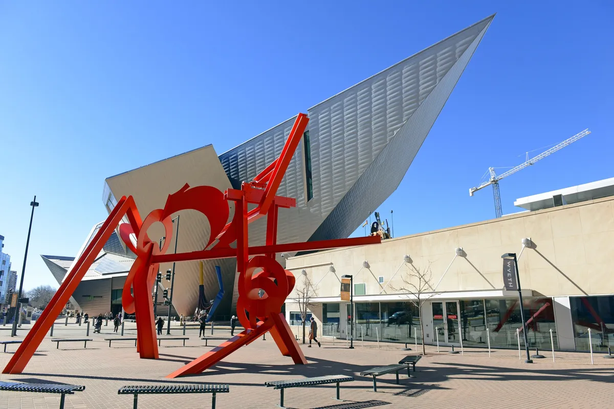 A striking red sculpture commands attention in front of a modern architectural building, creating a lively urban scene. The area is abuzz with visitors appreciating public art, taking photos, and engaging with the creative spirit of the city.