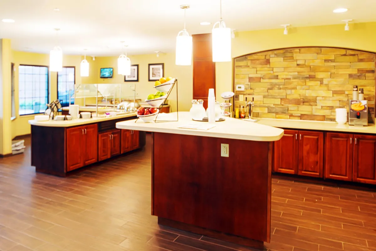 A spacious dining area featuring a central island with a countertop, a vibrant fruit display, stone wall accents, and warm lighting fixtures. The layout is designed for both meal preparation and socializing, with ample space for enjoying breakfast, snacks, or shared dinners.