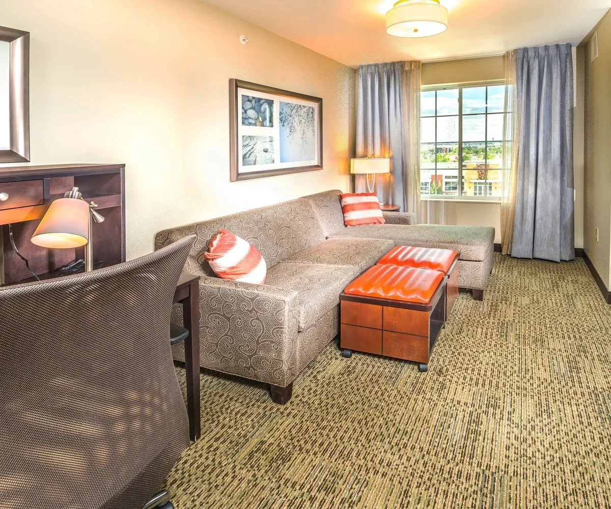 The image showcases a stylish, sunlit suite interior featuring a comfortable sofa, a modern coffee table, and a functional desk by the window. The space is designed for relaxation, reading, or remote work, with inviting natural light creating a warm, cozy atmosphere perfect for winding down or connecting with friends.