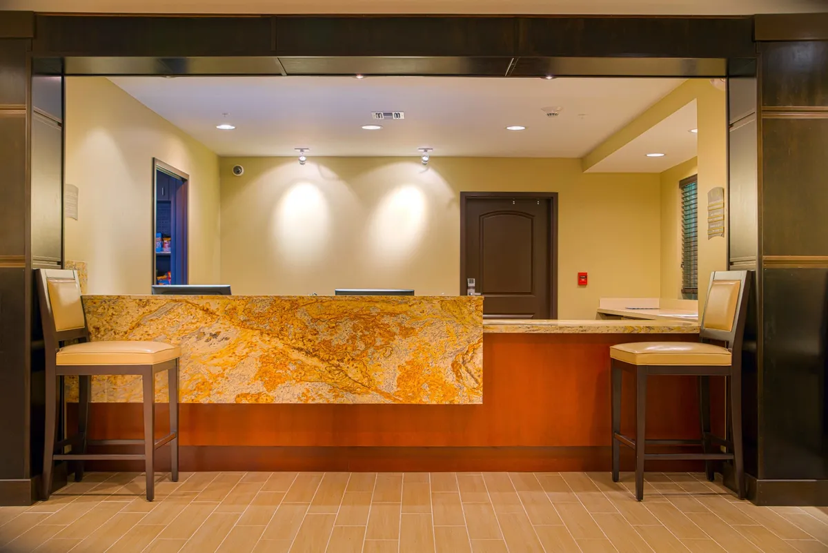 An inviting hotel lobby with a sleek granite reception desk, modern seating arrangements, and soft, ambient lighting. Staff members greet arriving guests with a warm welcome, while others relax or wait comfortably in the seating area, reflecting the hospitality and guest services provided.