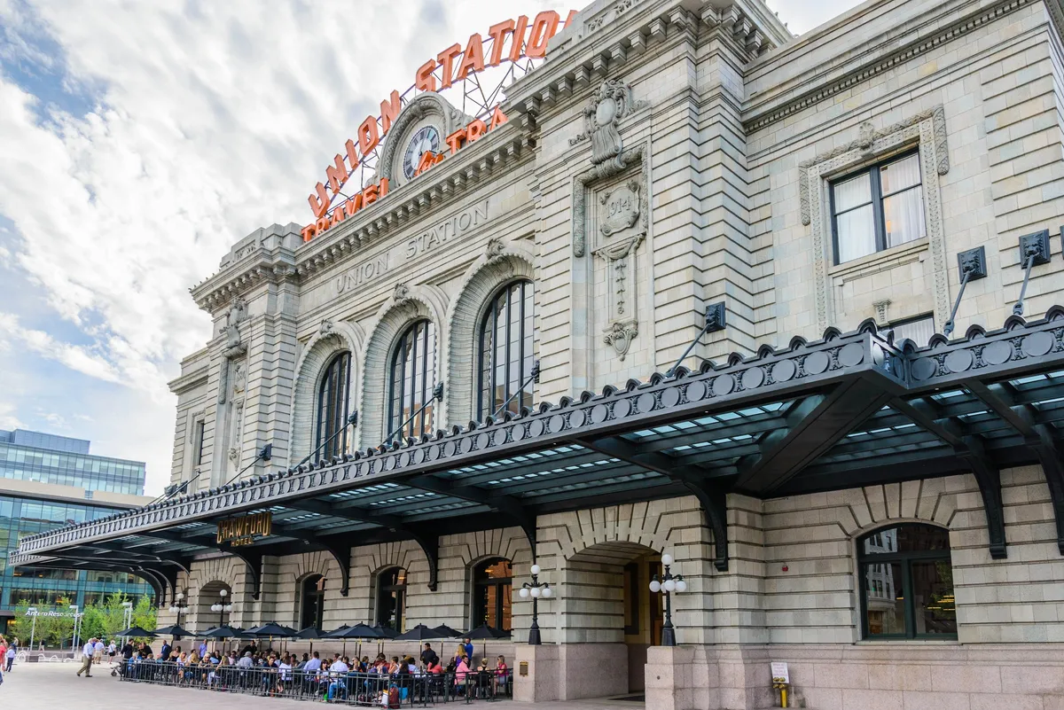 This image depicts the bustling exterior of Denver's iconic Union Station. Visible are the station's signature arched windows, the prominent 'Union Station' sign, decorative columns, and an active outdoor seating area filled with people. The scene captures cafes and passersby, conveying both movement and the charm of Denver's urban core.